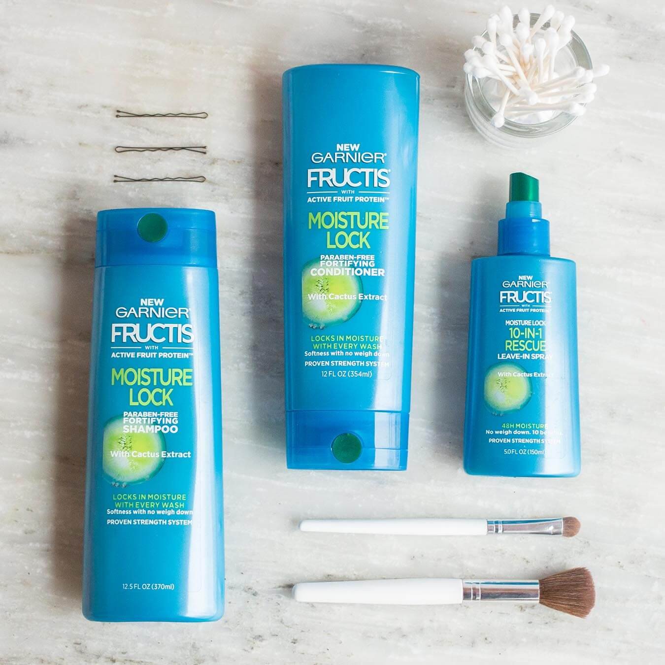 Garnier Fructis Moisture Lock Shampoo, Fructis Moisture Lock Conditioner, and 10-in-1 Rescue Leave-In Spray on grey marble next to three bobby pins, two makeup brushes, and a bowl of q-tips.